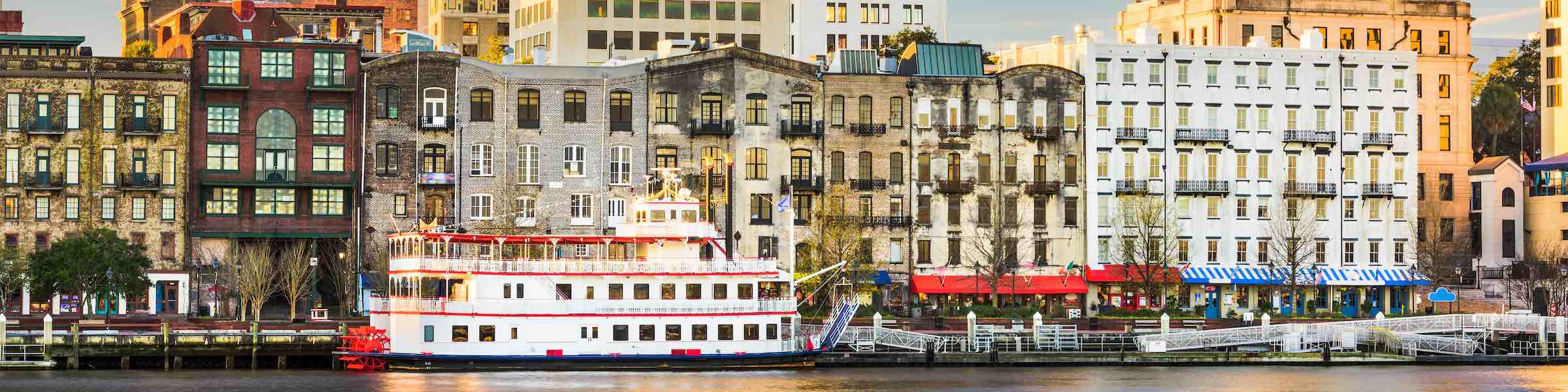 View of River Street, Savannah, GA, showing a red and white paddleboat in front of historic buildings formerly used as warehouses and offices for the shipping industry.