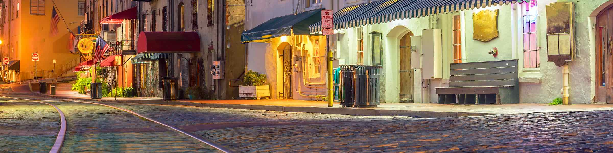 Night view of streetcar tracks on Savannah, GA's cobbled River Street, with illuminated shops and restaurants.